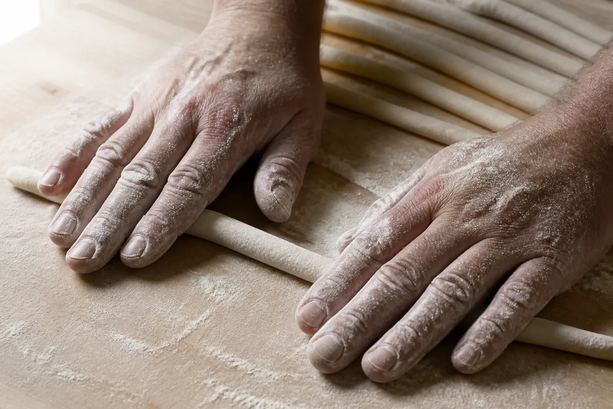 Mains farinées roulant un boudin de pâte sur un plan de travail en bois clair, lumière naturelle de fenêtre